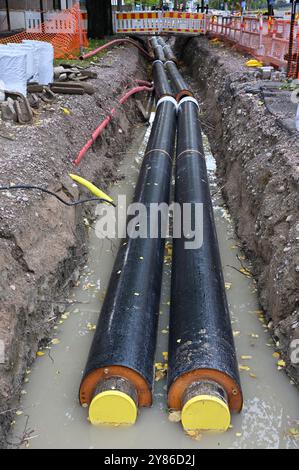 The image showcases a construction site focused on installing large underground pipes. Two large insulated pipes are visible in a trench filled with w Stock Photo