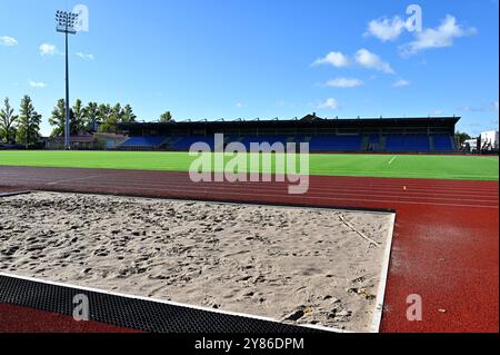 This image depicts an athletic stadium under clear blue skies, showcasing a long jump sandpit in the foreground, a red running track, and a vibrant gr Stock Photo