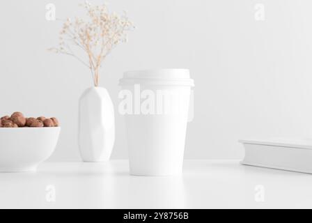 Coffee paper cup mockup with a gypsophila in a vase, hazelnuts and a book on a white table. Stock Photo