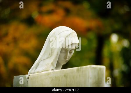 behind a white gravestone, a cracked female head of an also white stone statue peers out in front of colorful autumn leaves in blurred background Stock Photo