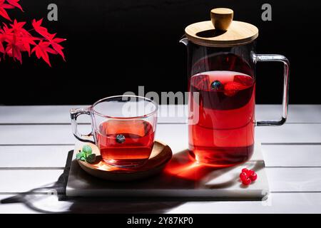 Red fruit infusion in crystal glass and pitcher on white wooden table on dark background. Stock Photo