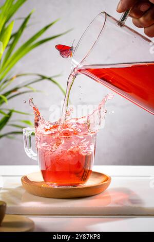 Pouring red fruit infusion and splashing from pitcher into glass on light background. Vertical format. Stock Photo