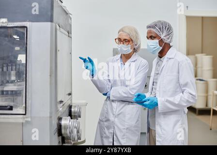 Medium shot of middle aged female process technician in lab coat pointing at machine, while inspecting drug production with African American male colleague at pharmaceutical manufacturing plant Stock Photo
