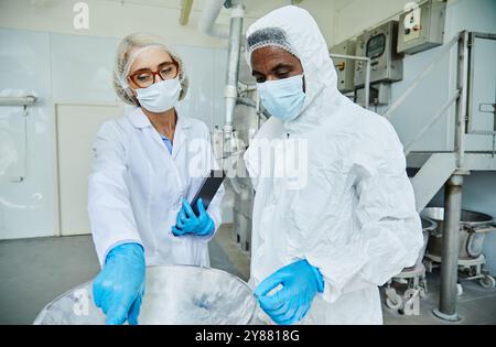 Medium shot of female process supervisor in lab coat pointing at equipment controlling production quality with African American male manufacturing technician, working in protective suit at factory Stock Photo