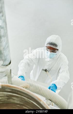 Vertical shot of African American male technician in blue gloves and disposable coveralls working with factory equipment on production floor of pharmaceutical manufacturing plant, copy space Stock Photo