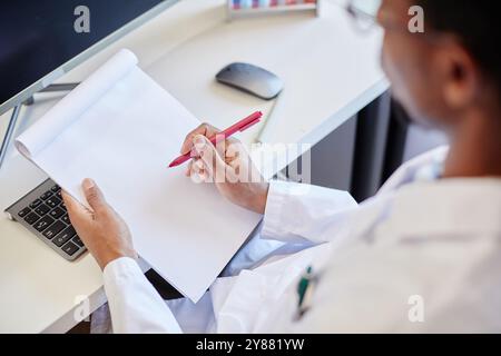 Over shoulder shot of African American male doctor or laboratory scientist making records in notepad while sitting at working table equipped with computer, copy space Stock Photo