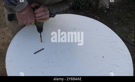 drilling holes in round white table top with screwdriver, top view, attaching table top to base, making DIY outdoor furniture Stock Photo