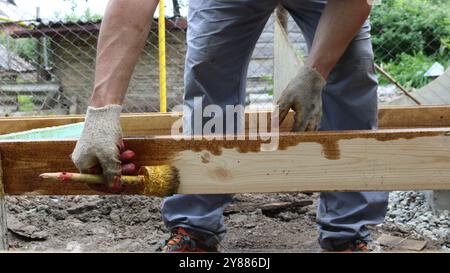 In the backyard a construction man in gray work pants and work gloves is painting a wooden beam. In his hand he holds a brush covered with varnish Stock Photo