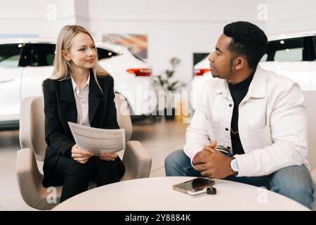 Portrait of female car dealer consulting doubtful black buyer, handing over purchase agreement for review at showroom. Serious African client Stock Photo