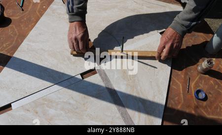 A man is holding a wooden bar or ruler used for marking or measuring on ceramic tiles with a marble pattern. A diagonal line for cutting is drawn Stock Photo