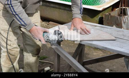 A man in work clothes is using a hand-held electric tool to polish a ceramic tile with a marble pattern that lies on a wooden table Stock Photo