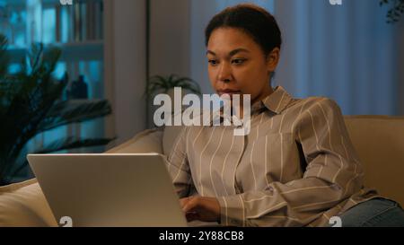 Focused African American woman ethnic girl 30s female working with laptop on comfortable couch use computer device on sofa browsing internet social Stock Photo