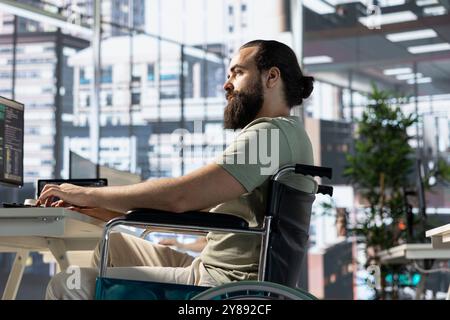 IT employee with disability sitting in wheelchair in office working at computer desks, testing and deploying programs and systems. Programmer with paraplegia in inclusive workplace checking code on PC Stock Photo