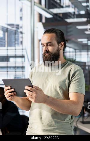 Man working in office developing software in Java programming language using device. Programmer walking through workspace with tablet, thinking how to solve coding problems Stock Photo