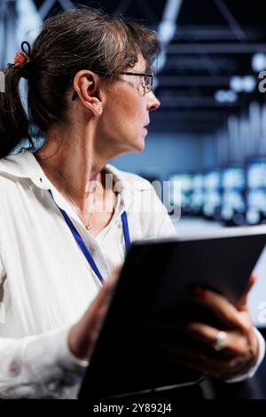 Old computer scientist updating data center rackmounts, writing intricate binary code scripts on tablet, using programming to fortify cybersecurity in technically advanced supercomputer hub Stock Photo