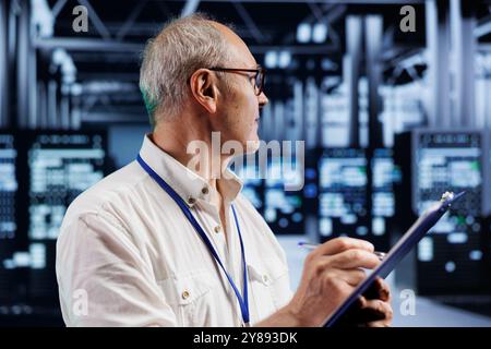 Elderly engineer in crypto mining farm with specialized hardware using Blockchain technology. Fintech expert holding clipboard, inspecting asic rigs solving complex computational problems Stock Photo