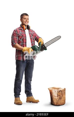 Full length portrait of a young man holding a chainsaw by a tree trunk isolated on blue background Stock Photo
