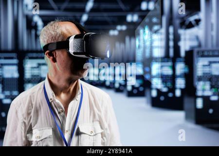 Old cloud computing business executive immersed in virtual reality at data center, doing equipment maintenance. Repairman using VR headset to optimize server rigs performance, checking code lines Stock Photo