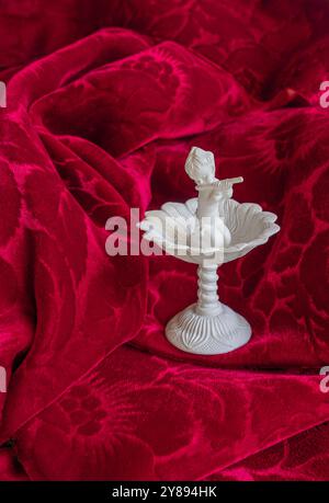 Red fabric background is velvet. Musician musical performer figure figurine plays the flute, Statue is in white on a pedestal. A recital is happening. Stock Photo