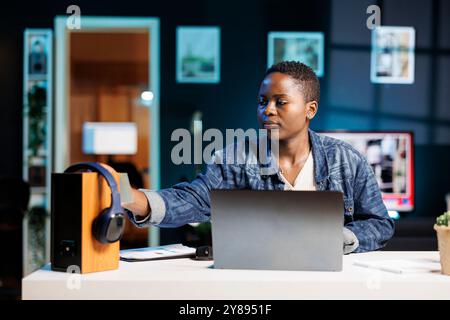 Young black woman working from home using a laptop and placing a sticky note on a speaker. African American blogger putting post-it notes on a device while using her personal computer. Stock Photo