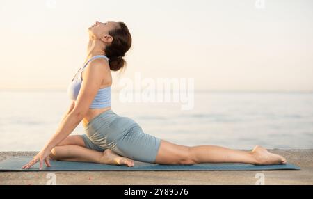 Active happy woman in sportswear sitting on yoga mat and doing stretching legs during yoga on seashore at sunrise Stock Photo