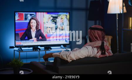 Arabic man lounged on cozy sofa, watching news broadcast while relaxing in apartment after hard day at work. Middle Eastern person chilling on couch, looking at TV channel showing current events Stock Photo