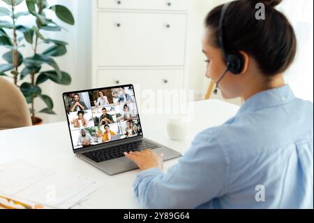 Side view to caucasian confident business woman with a headset using laptop to talk with colleagues, discussing business plan and strategy via video call while sit at a workplace. Online communication Stock Photo