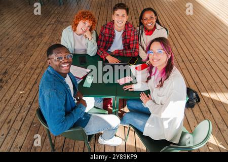 High angle view of diverse group of smiling students looking at camera while working together around a table, with notebooks and smartphones, studying and collaborating in a casual setting indoors. High quality photo Stock Photo