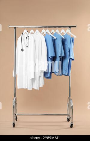 Different medical workers' uniforms and stethoscope on clothing rack against beige background Stock Photo