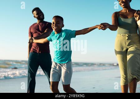 African american playful son holding father and mother hands and walking at shore against sky Stock Photo