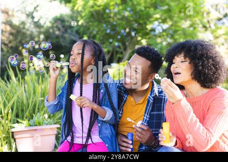 Happy african american father, mother and daughter blowing bubbles in sunny garden, copy space Stock Photo