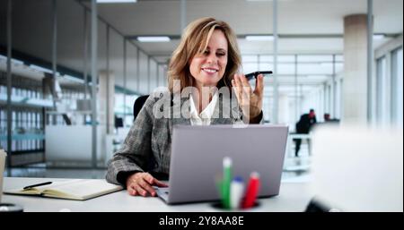 Professional Businesswoman Using Smartphone App For Client Call In Modern Office Setting With Laptop And Smiling Stock Photo