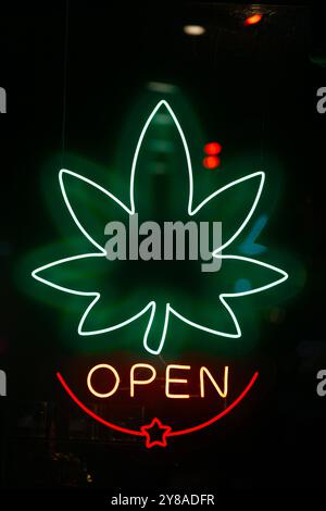 A neon sign in a store window shaped like a cannabis leaf with the word 'Open' written underneath. Stock Photo