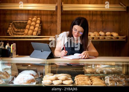 Baker at work recording orders in a cozy bakery surrounded by pastries Stock Photo