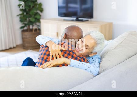 Happy diverse senior couple sitting on sofa and hugging in sunny living room Stock Photo