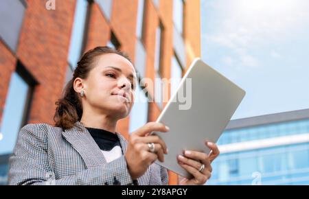Professional woman standing outdoors, using a digital tablet, with a modern building in the background Stock Photo