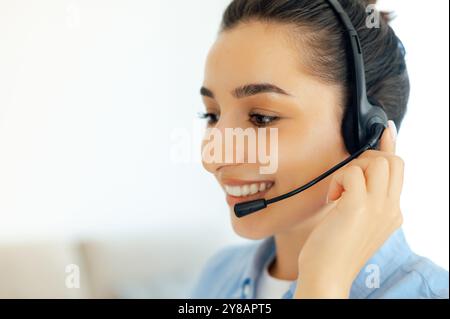 Side view close-up photo of a positive successful brunette caucasian woman with headset, working as call center operator, customer support service, hotline assistant, smiles while talking with client Stock Photo