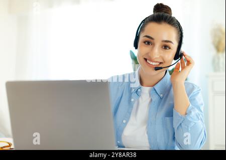 Well dressed charming successful caucasian woman with wireless headset, working in customer support service or helpline, female call center agent, sits at her work desk with laptop, smiles at camera Stock Photo