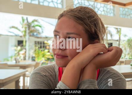 A young woman sits at a cafe table, resting her chin on her arms and gazing thoughtfully outside. Stock Photo