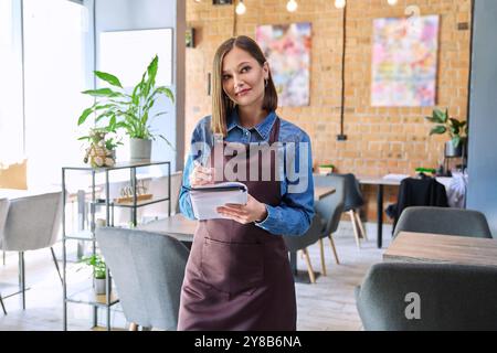 Young woman worker, owner in apron holding notepad pen in restaurant, coffee shop Stock Photo
