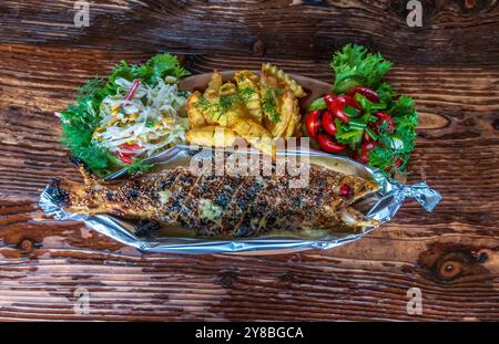 Close-up top view of freshly caught baked trout with sauce with sauce, french fries and fresh juicy salads on a wooden rustic table Stock Photo