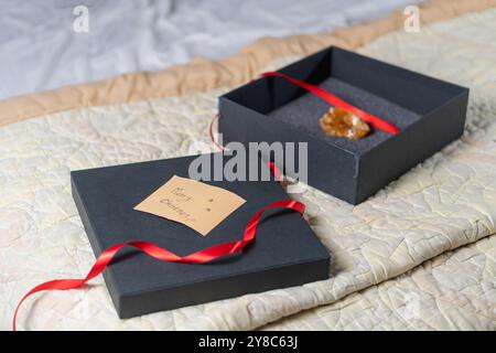 A black gift box with a red ribbon, partially open, revealing a small, shiny object inside. The box is placed on a textured, light-colored quilt. A no Stock Photo