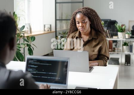 Young African American woman working in light spacious office with her unrecognizable colleague who writing code Stock Photo