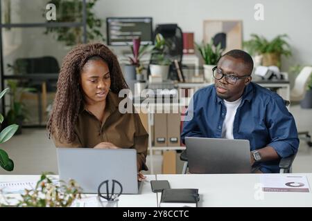 African American man holding silver laptop on knees helping his beautiful curly haired female colleague during shift in office Stock Photo