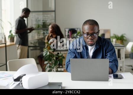 Young African American man with glasses and dressed in casual clothes working via silver laptop while his colleague chatting in blurred background Stock Photo