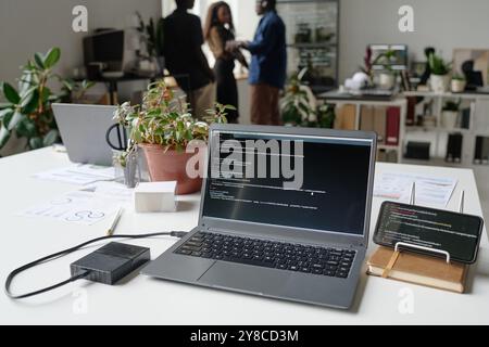 High angle shot of working silver laptop with codes on screen, unrecognizable programmers chatting in blurred background Stock Photo
