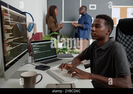 Young African American app developer sitting at white work desk with computer, coffee cup and laptop, he writing codes while his colleagues chatting Stock Photo