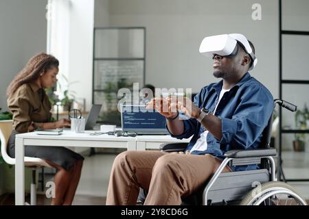 African American worker with disability gesticulating while playing VR game during break, his colleagues working in background Stock Photo
