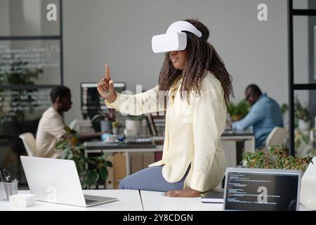 Curly haired woman playing video game while sitting on work table with laptops Stock Photo