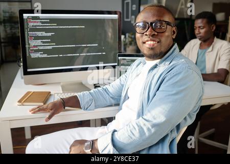 Medium closeup portrait of joyful programmer sitting at table and looking at camera Stock Photo